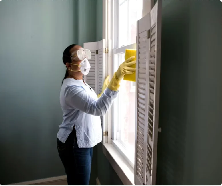 Woman wearing protective goggles, face mask, and yellow gloves while cleaning a window with a yellow cloth.